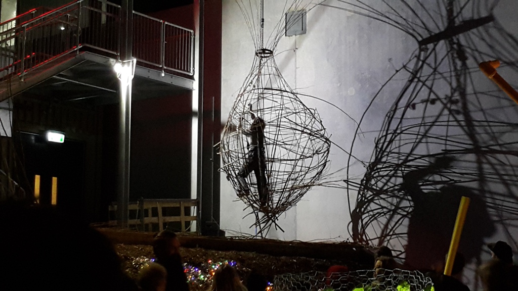Sculpture artist suspended above a crowd at nighttime, weaving a wire basket around herself, which casts a shadow against a concrete wall next to her