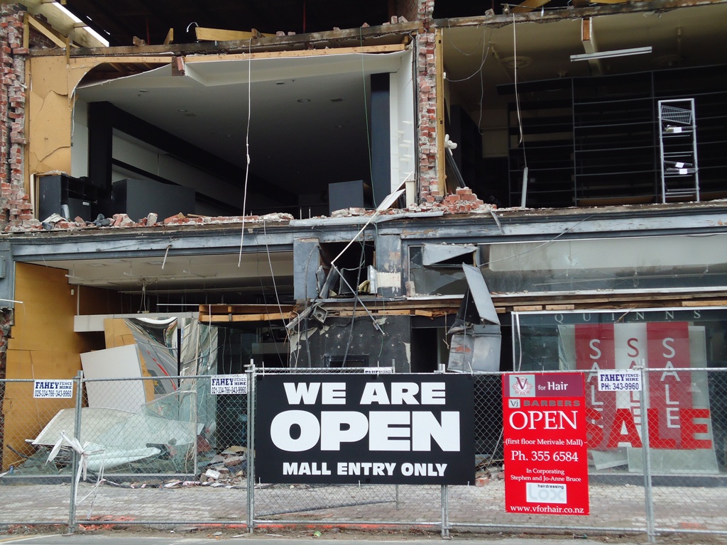 Photo of a sign in front of a severely earthquake damaged building stating 'Merivale Mall. We are open' also showing an arrow to alternative entrance
