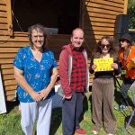Three people standing smiling at a camera, one woman proudly holding a yellow certificate up to the camera