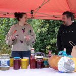 Woman and man chatting animatedly as they stand behind a trestle table laden with preserve jars with a variety of different coloured preserves