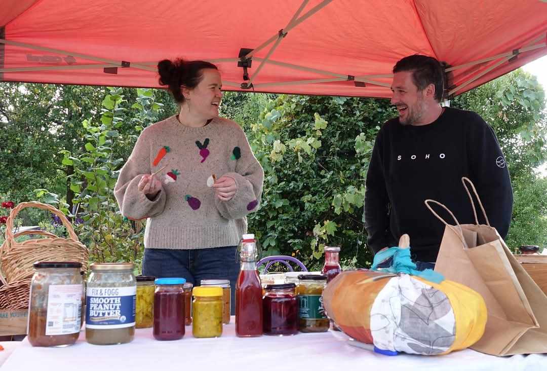 Woman and man chatting animatedly as they stand behind a trestle table laden with preserve jars with a variety of different coloured preserves